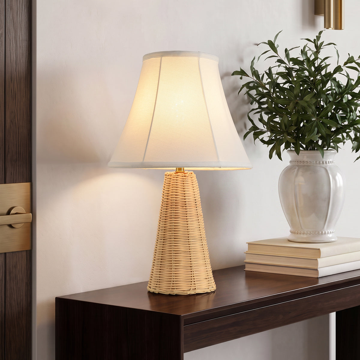 Woven rattan table lamp with a white shade on a dark wooden console table next to a white vase.